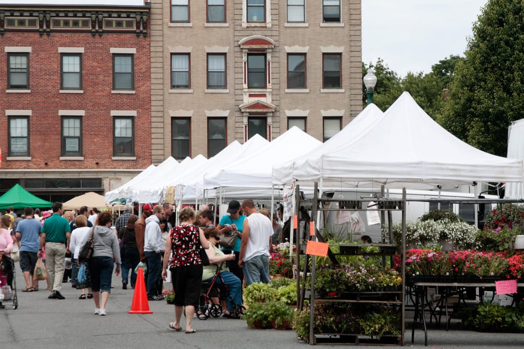 Groups of people shop at white pop-up tents full of produce and flowers at a farmer's market.