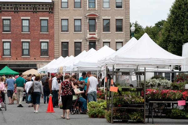 Groups of people shop at white pop-up tents full of produce and flowers at a farmer's market.